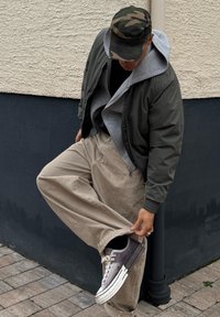 Personne portant une casquette camouflage, une veste verte et un pantalon beige, ajustant la jambe du pantalon pour montrer une chaussette noire et une chaussure grise à un coin de trottoir.