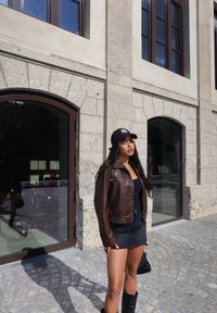 Καφέ δερμάτινο μπουφάν με φερμουάρ, μαύρη μπλούζα tank top και τζιν φούστα; μαύρο καπέλο μπέιζμπολ; μαύρη τσάντα; πέτρινη επιφάνεια και λιθόκτιστο κτίριο.