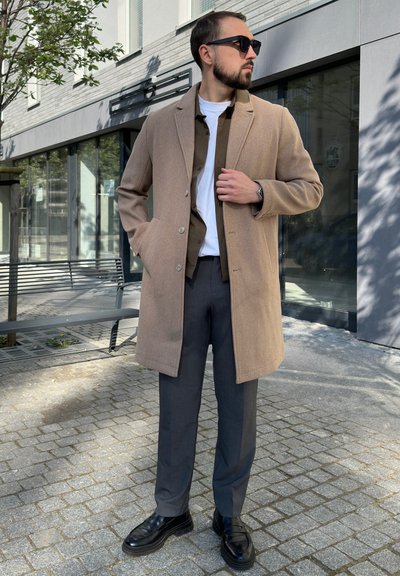 Abrigo de lana beige sobre una camisa marrón oscura, combinado con pantalones grises y zapatos negros. Las gafas de sol completan el atuendo contra un fondo urbano.