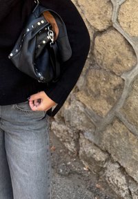 Fekete bőr kézitáska ezüst szegecsekkel, egy fogantyúval és kapoccsal. Fekete pulóverrel és szürke farmerrel viselve, egy kőfal előtt.