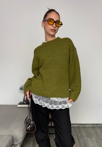 Olíva zöld, textúrált kötött pulóver fehér csipkés felsőre rétegezve. Fekete, laza szabású nadrág és egy kis sötét kézitáska tartozik hozzá.