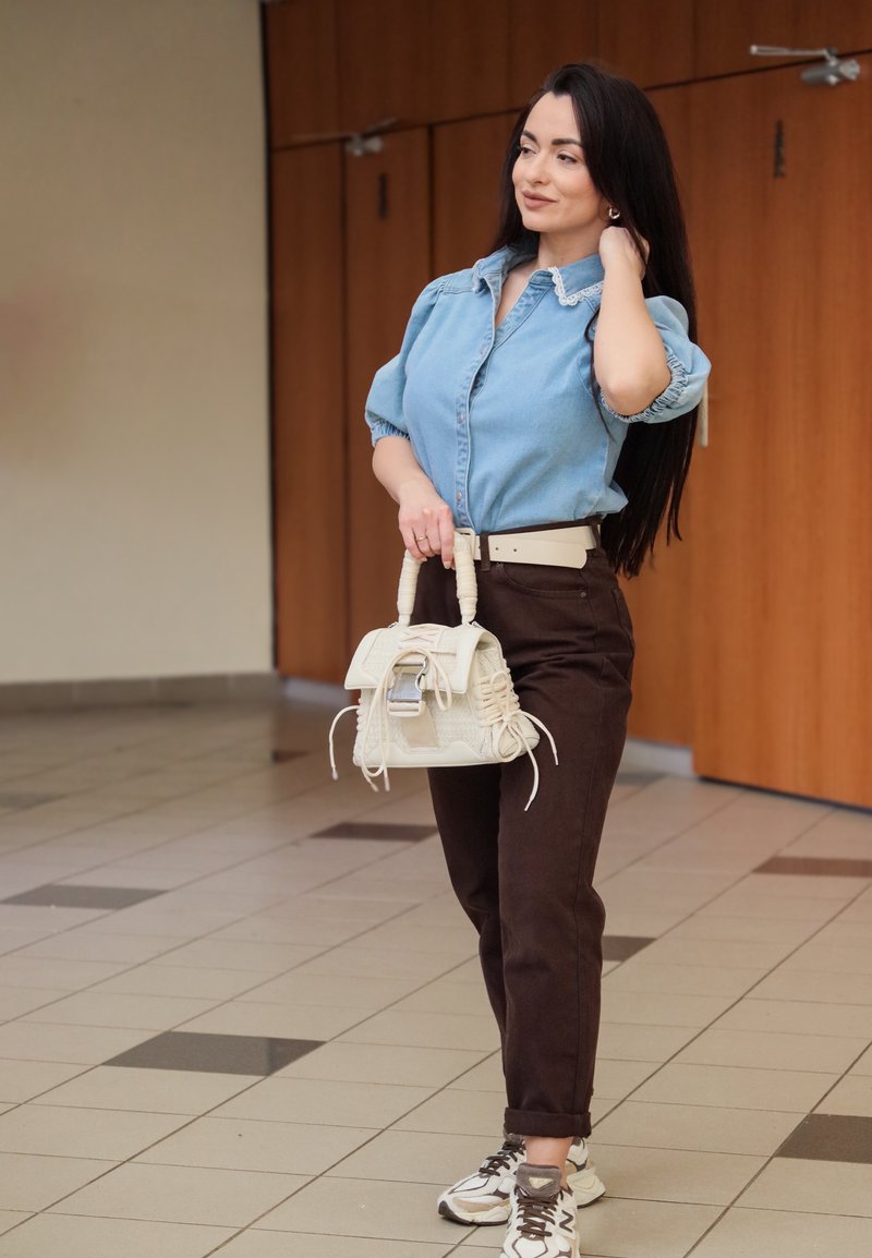 Camicia di denim con maniche a sbuffo, pantaloni a vita alta marrone scuro, cintura beige e borsa crema con dettagli testurizzati. Sneakers neutre.