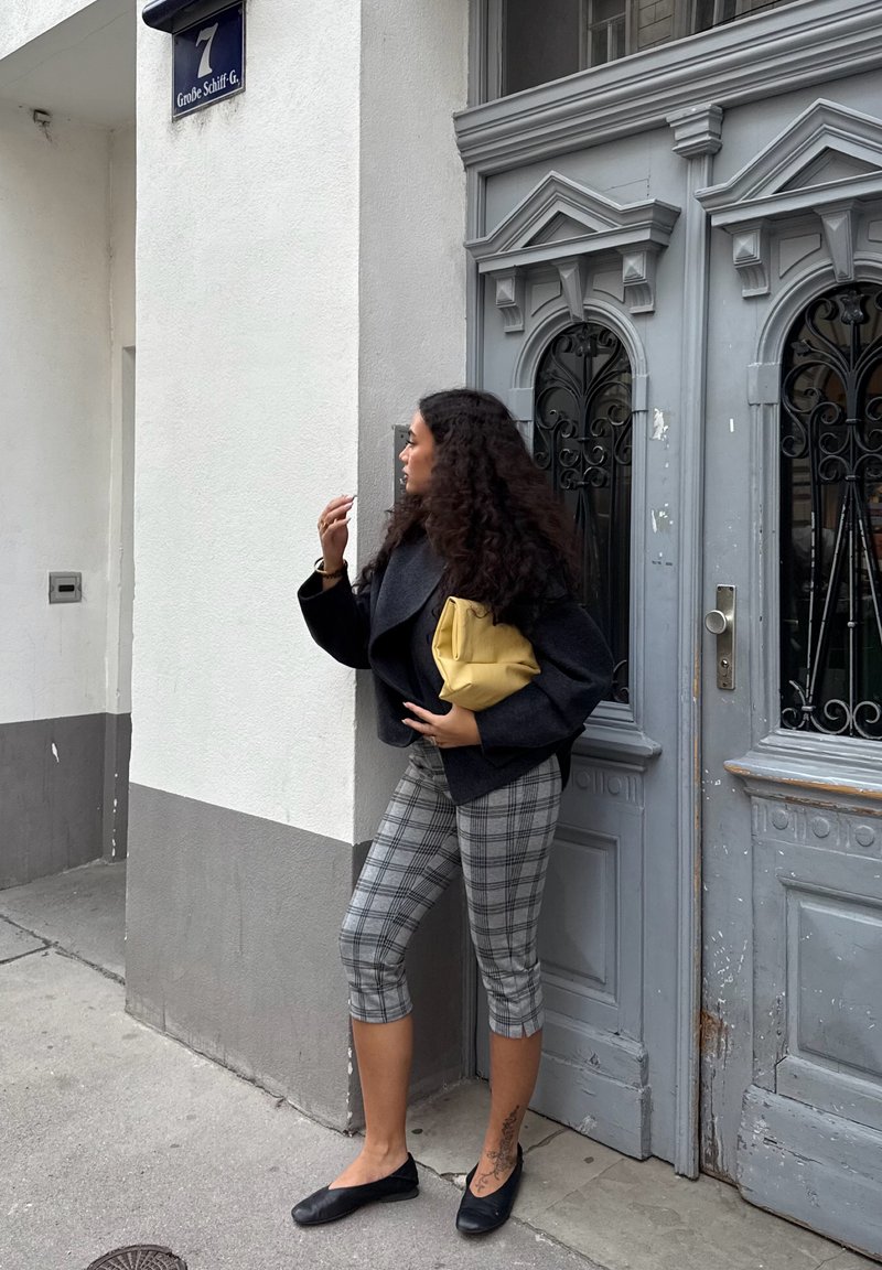 A woman dressed in a black cropped coat and plaid capri trousers, holding a yellow bag, stands beside an ornate grey door featuring black accents.