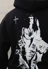 Svart hettegenser med en stor hvit illustrasjon av en kvinne med langt hår, som bærer et smykke, foran en teksturert bakgrunn. Akkompagnert av hvit tekst.