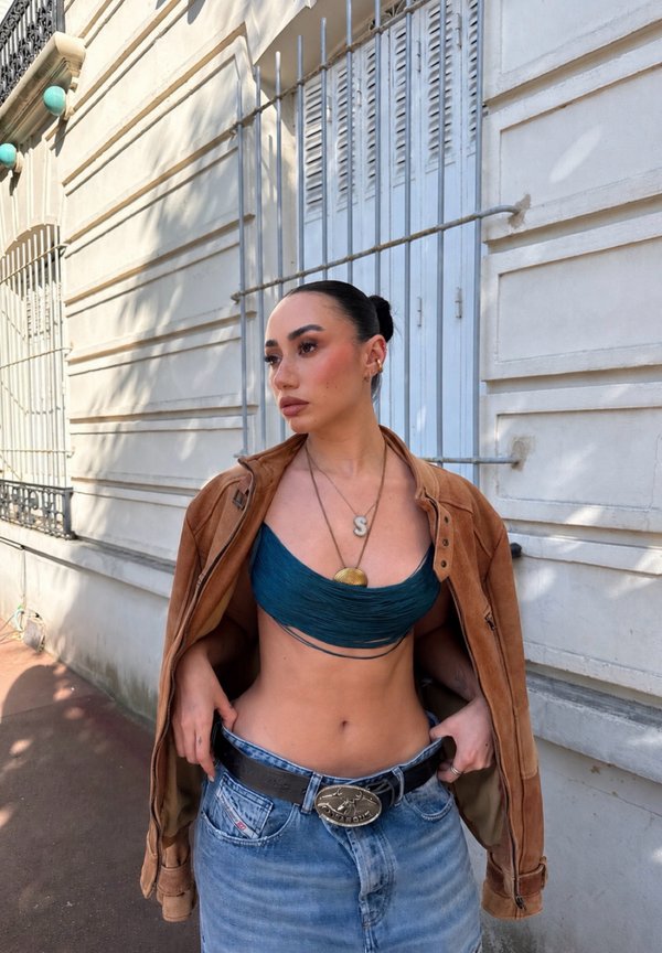 Femme aux cheveux foncés en chignon porte une veste marron sur un crop top bleu et un jean taille haute, debout contre un bâtiment en pierre claire avec des barreaux en métal.