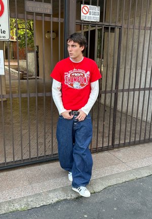 Joven con una camiseta roja de campeones de Ohio State Buckeyes y jeans holgados, de pie frente a una puerta de hierro cerrada con señales italianas de prohibido estacionar.