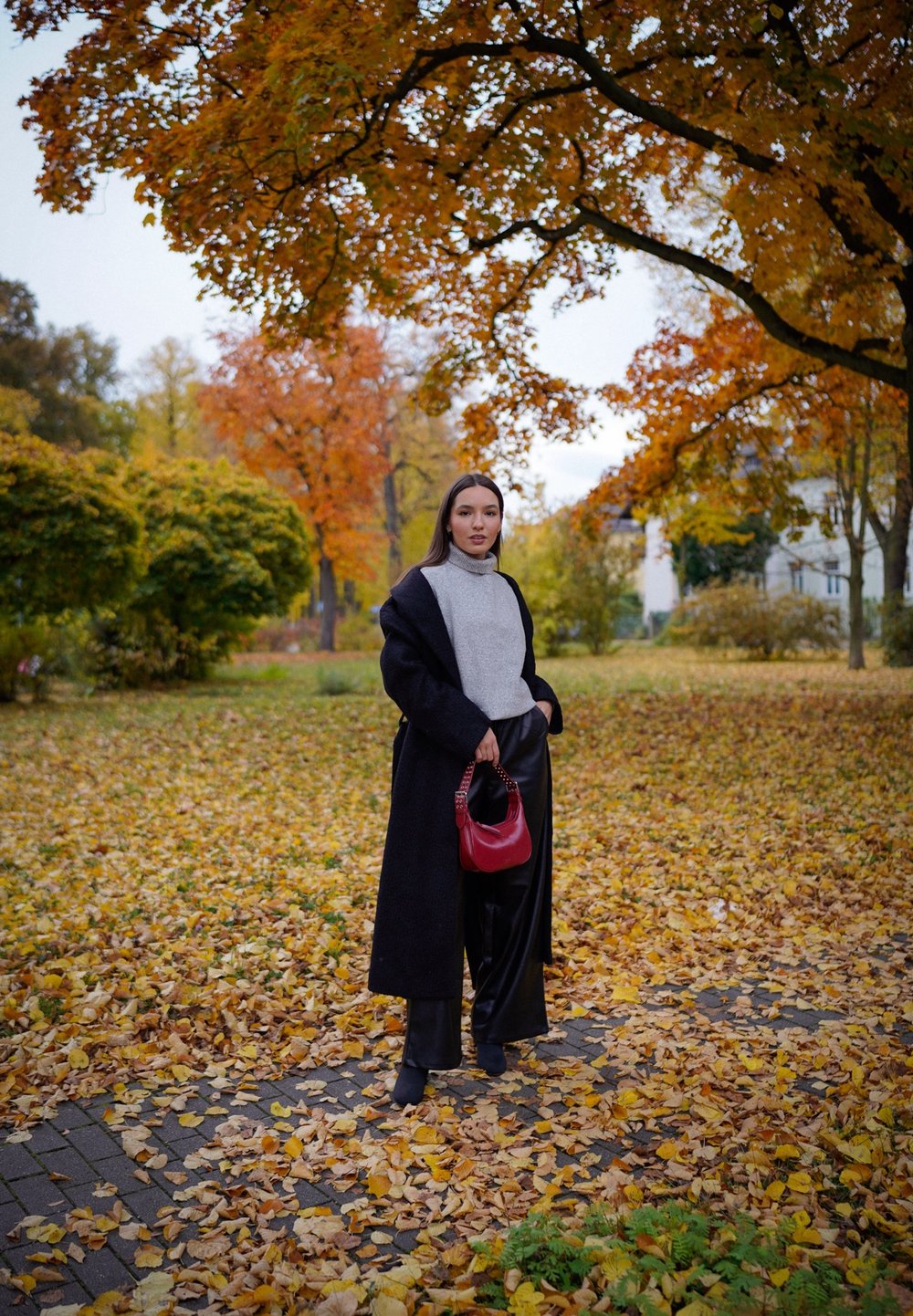 Casaco preto, suéter cinza, calças largas pretas; segurando uma bolsa vermelha. O fundo apresenta folhas de outono e árvores.