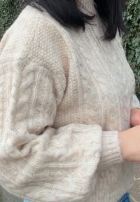 Femme portant un pull en maille torsadée couleur crème, debout à l'extérieur à côté de plantes vertes feuillues.