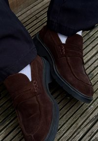 Scarpe in suede marrone con punta rotonda e doppia cinghia, appoggiate su una superficie di legno, abbinate a calzini bianchi e pantaloni scuri.