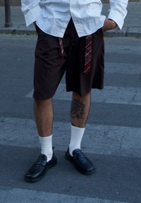 Rudos šortai su laisvu kirpimu, vilkimi su balta marškiniais, juodais loaferiais ir ilgomis baltomis kojinėmis. Ant kojos matomas tatuiruotė.