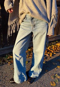 Jean large bleu clair en denim, associé à un pull gris avec des accents tricotés et des franges aux manches. Chaussures noires visibles.