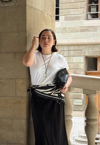 Camicia bianca a maniche corte, gonna nera, tessuto a motivo zebra in vita, tiene una borsa nera, in piedi vicino a una colonna di pietra in uno spazio luminoso.