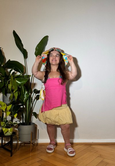 Top sin mangas de color rosa brillante, falda plisada amarilla clara, sombrero de ala ancha con estampado, sandalias blancas. Plantas verdes en el fondo.