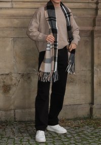 Pull en tricot beige à coupe ample, pantalon noir et écharpe à carreaux beige, noir et blanc, assortis à des baskets blanches.