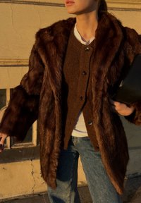 Brun faux fur jakke med bred krave, tekstureret finish og store knapper, lagd over en brun strik cardigan og en hvid skjorte, parret med denim.