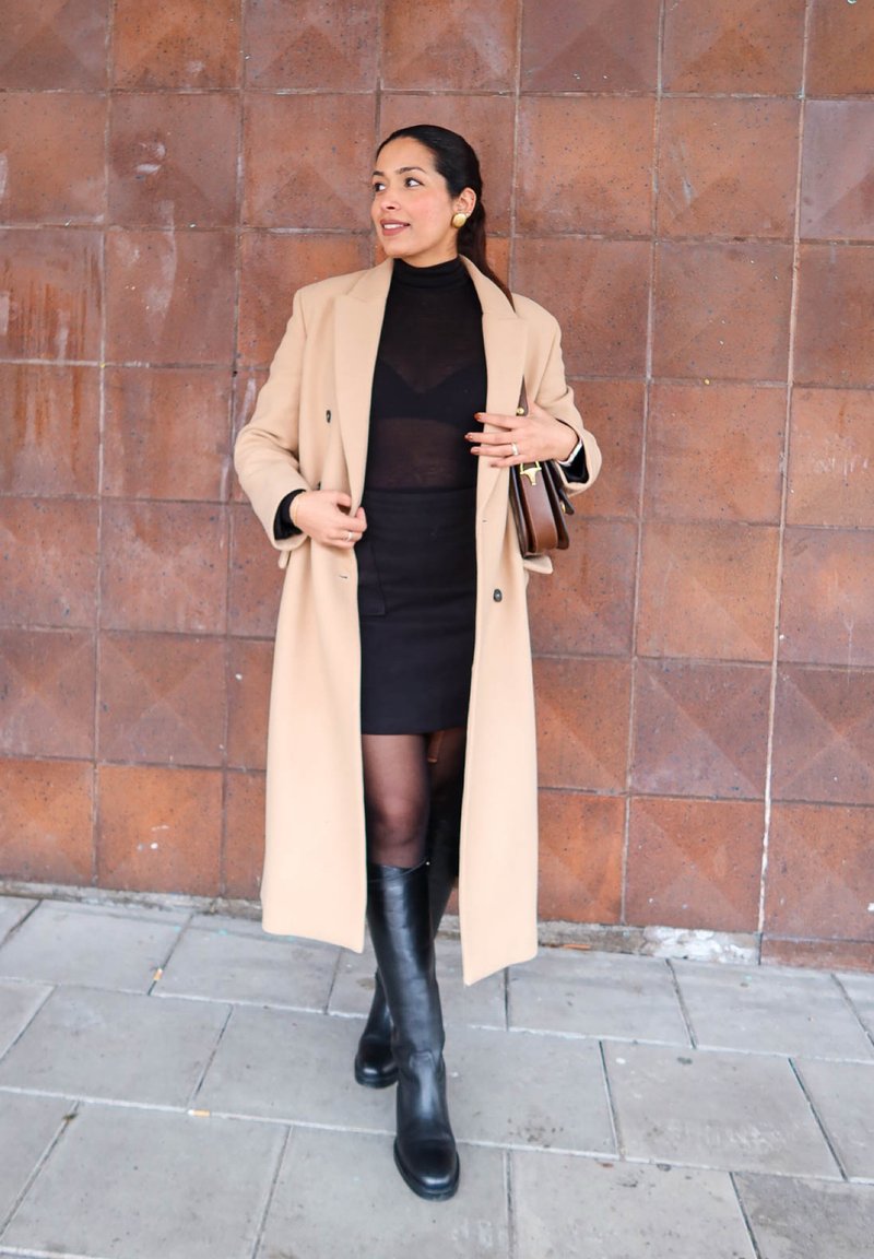 Mujer con abrigo largo beige, vestido negro, botas hasta la rodilla y un bolso marrón en la mano, de pie frente a una pared de azulejos.