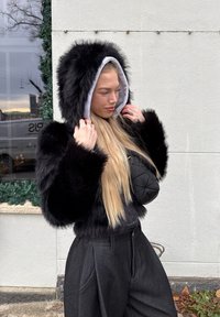 Jachetă scurtă din blană artificială neagră cu mâneci groase și pufoase, căptușeală cu glugă albă și asortată cu pantaloni înalți, închiși la culoare.