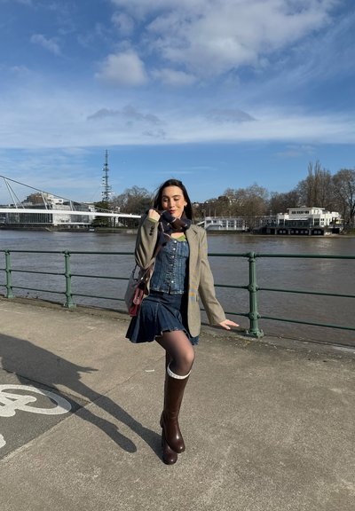 Mujer posando en un paseo junto al río, vistiendo un vestido de mezclilla, blazer, bufanda y botas hasta la rodilla bajo un cielo azul con nubes dispersas.