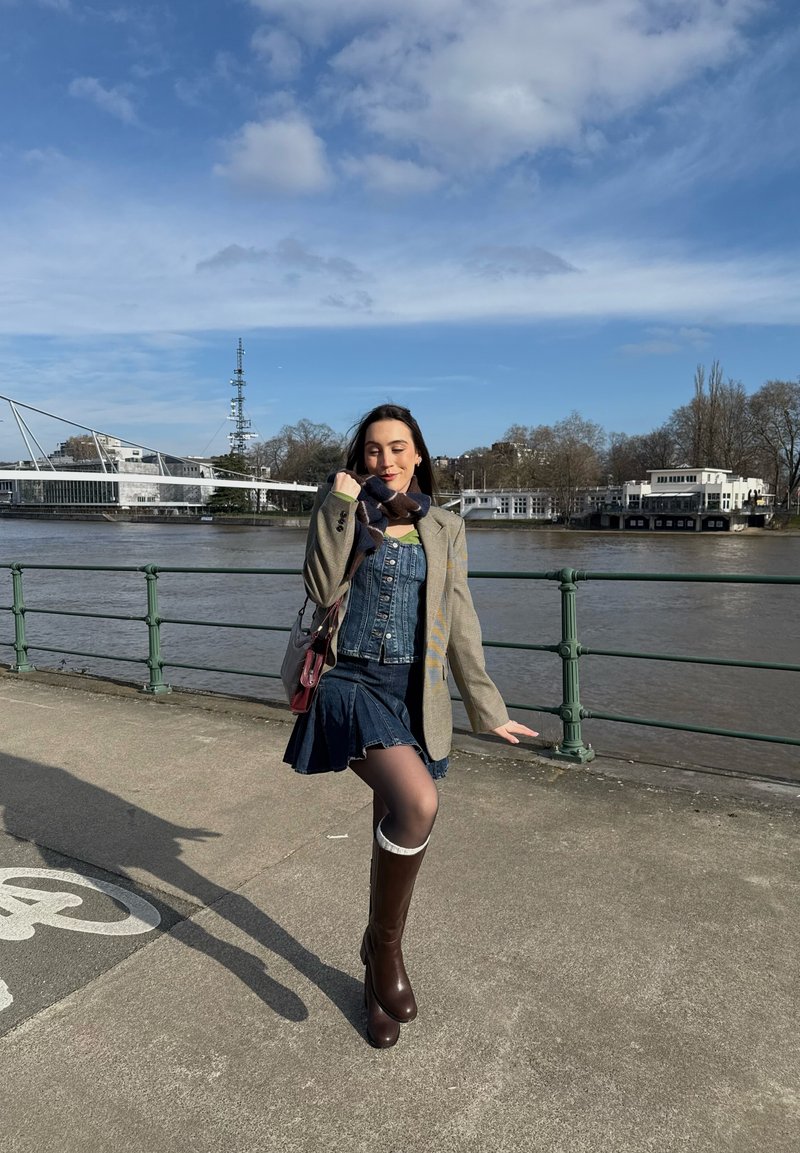 Nő pózol a folyóparti sétányon, farmerruhában, blézerben, sállal és térdig érő csizmában, kék ég alatt, apró felhőkkel.