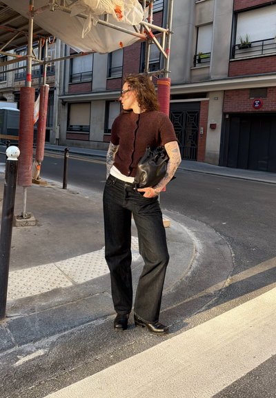 Mujer con tatuajes que lleva pantalones oscuros y una blusa marrón, sosteniendo una bolsa negra, de pie en una calle urbana cerca de andamios a la luz del día.