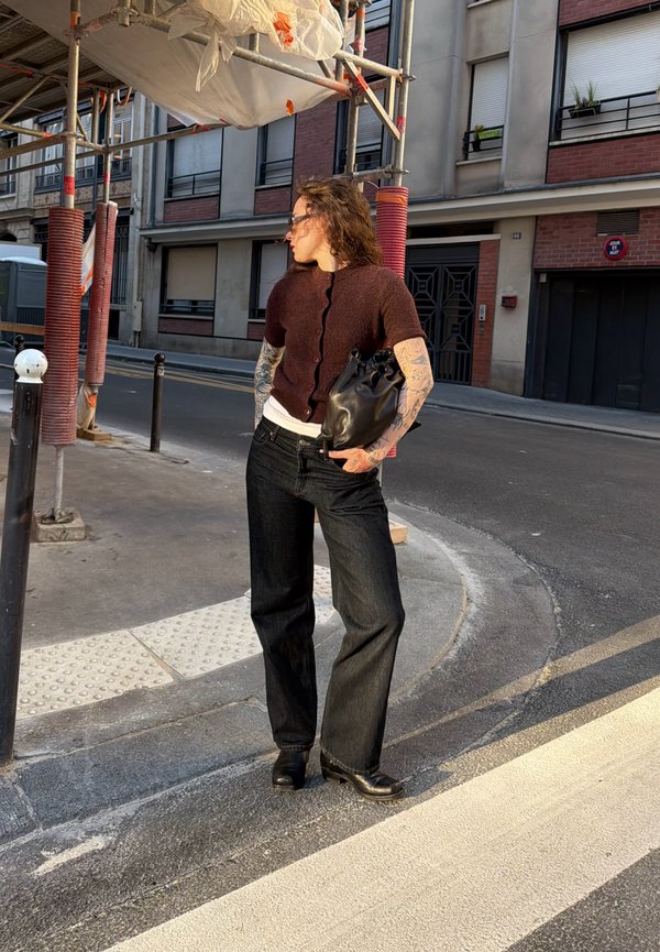 Woman with tattoos wearing dark pants and brown top, holding black bag, standing on urban street near scaffolding in daylight.
