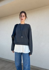 Sivi oversized pulover s rebrastim rubom, složen preko bijele svilenkaste bluze, u kombinaciji s plavim širokim trapericama. Uključuje crnu torbu preko ramena i ogrlicu.