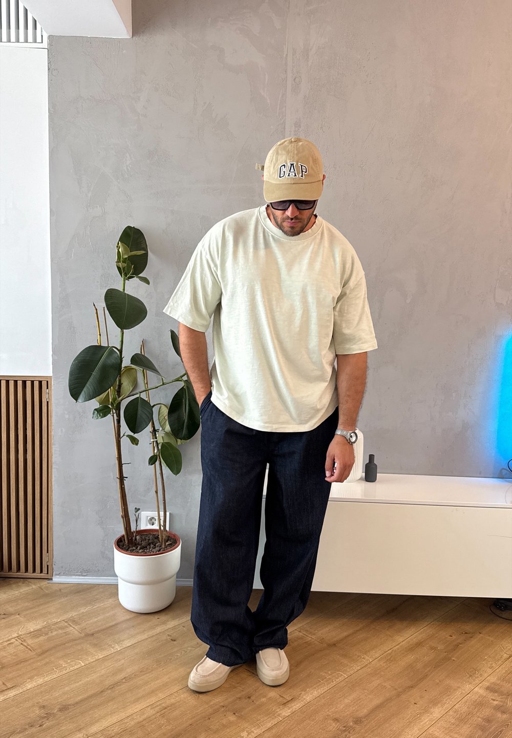 Light green textured T-shirt, dark blue wide-leg jeans, beige cap with "GAP" logo, light beige shoes, standing beside a potted plant.