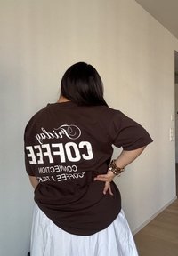 Osoba z długimi ciemnymi włosami, ubrana w brązową koszulę z napisem "Friday COFFEE CONNECTION" oraz białą spódnicę, stojąca w pomieszczeniu, przodem do ściany.