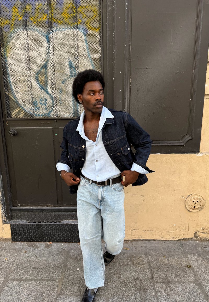 Hombre con peinado afro que lleva una chaqueta vaquera, camisa azul claro, jeans y zapatos negros, de pie en una calle pavimentada cerca de una puerta cubierta de grafitis.