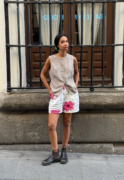 Chaleco sin mangas de canalé en tono marrón con botones frontales, combinado con pantalones cortos blancos con grandes patrones florales en color rosa; botas negras hasta el tobillo.