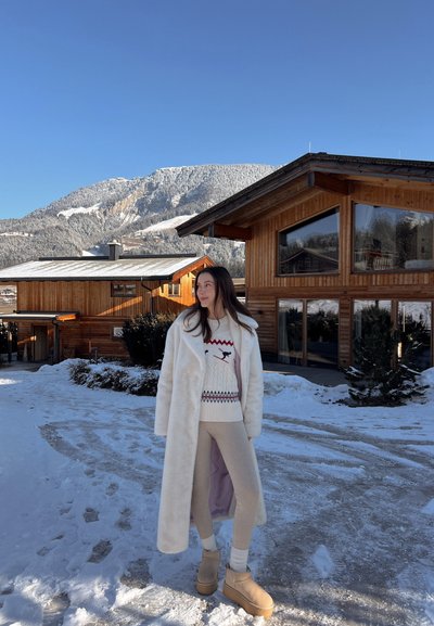 Abrigo de piel sintética blanca, suéter de punto crema con patrones, leggings beige y botas marrón claro, con un fondo de montaña nevada y edificios de madera.