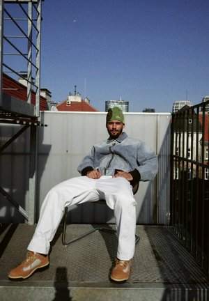 A denim jacket in a grey tone, white trousers, and brown shoes. A green beanie adds a pop of colour. Sitting on a metal chair with a rooftop backdrop.