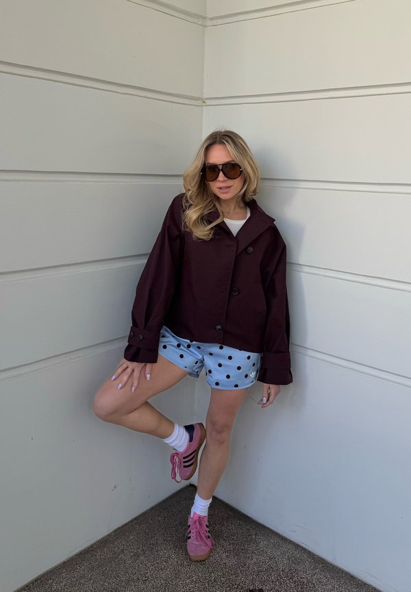 Frau mit Sonnenbrille, dunklem, oversized Jacke, hellblauen gepunkteten Shorts, weißen Socken und rosa Sneakers, die sich an eine weiße Wandlehne lehnt.