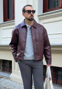 Bărbat îmbrăcat cu ochelari de soare negri, jachetă din piele maro, pulover gri și pantaloni gri, ținând o geantă de umăr bej lângă fațada unei clădiri.