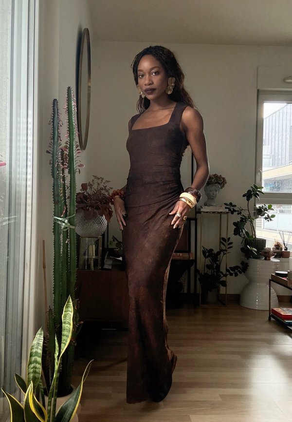 Femme aux longs cheveux tressés se tenant à l'intérieur, vêtue d'une robe marron sans manches et longue jusqu'au sol, portant plusieurs bracelets sur son bras gauche.