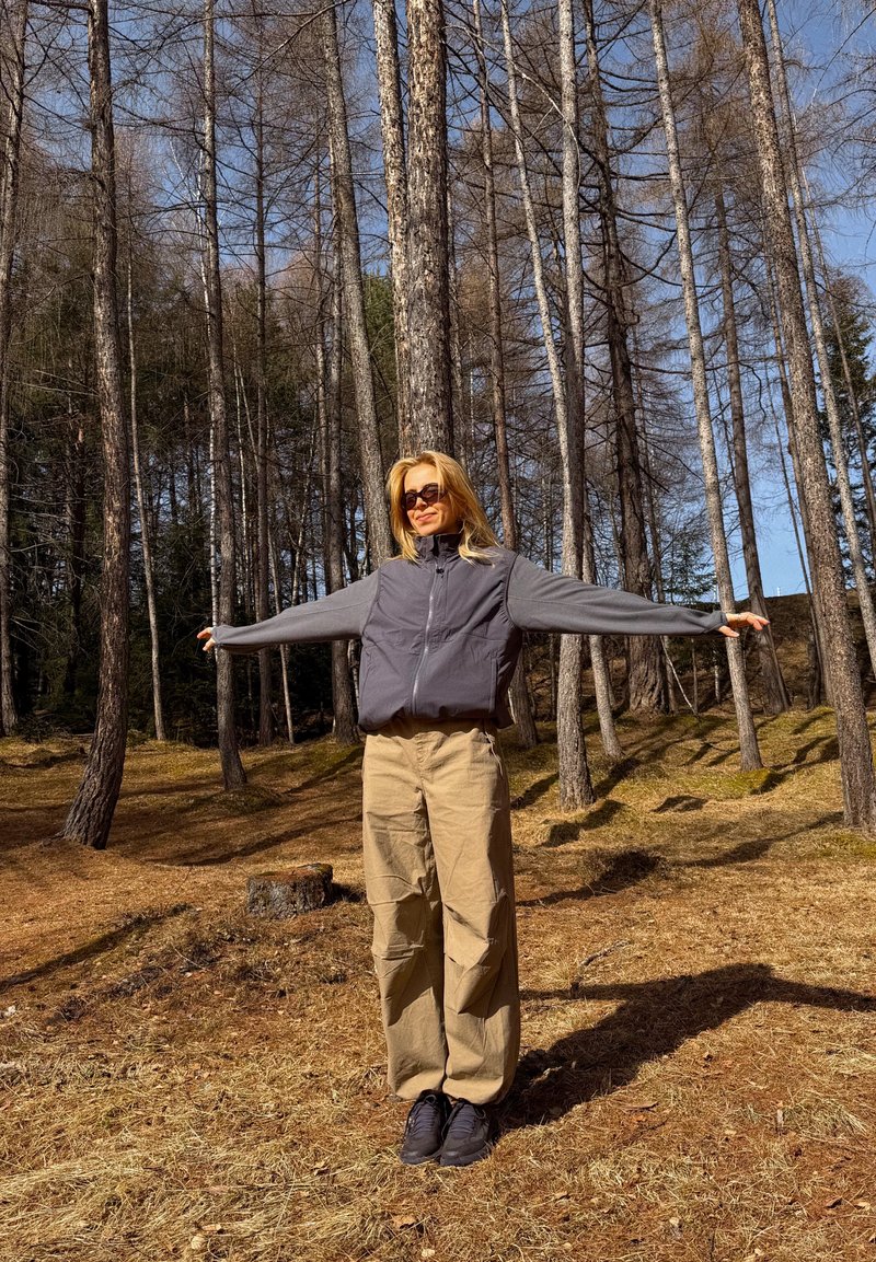 Frau mit Sonnenbrille, grauer Jacke und beiger Hose steht mit ausgestreckten Armen in einem sonnenbeschienenen Wald mit hohen, kahlen Bäumen.