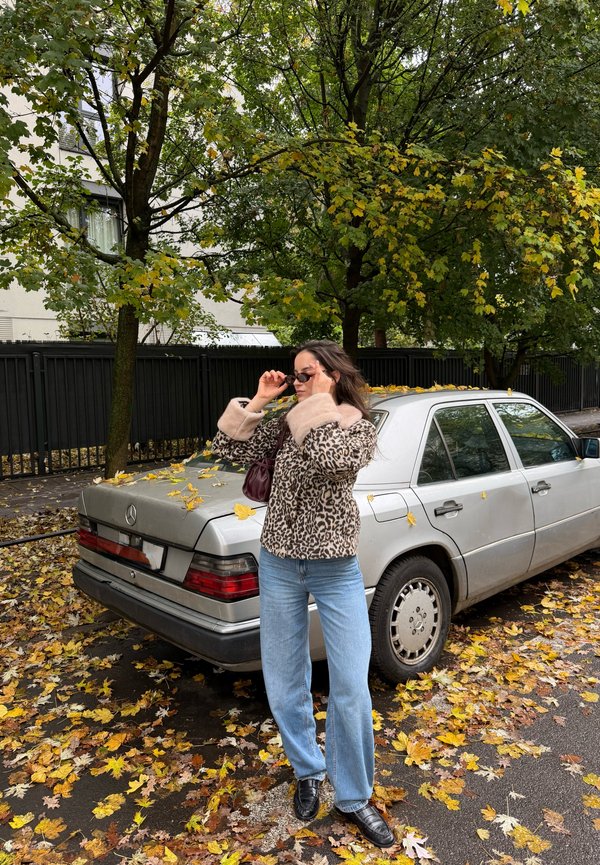 Une femme ajuste ses lunettes de soleil à côté d’une Mercedes argentée garée sous des arbres aux couleurs automnales, tandis que des feuilles mortes recouvrent la rue.