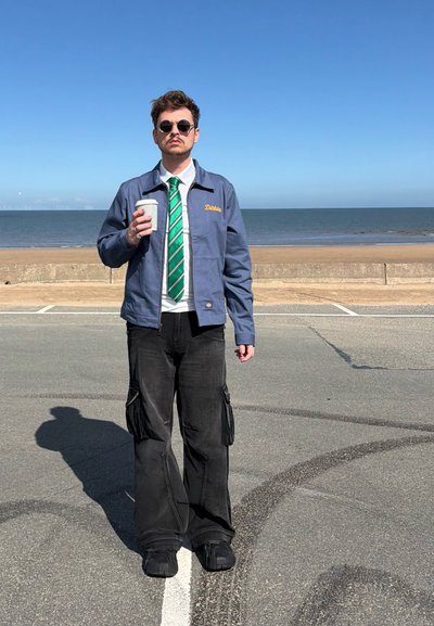 Hombre con gafas de sol, corbata verde a rayas y chaqueta azul sosteniendo una taza de café, de pie en una carretera cerca de la playa con el océano al fondo.