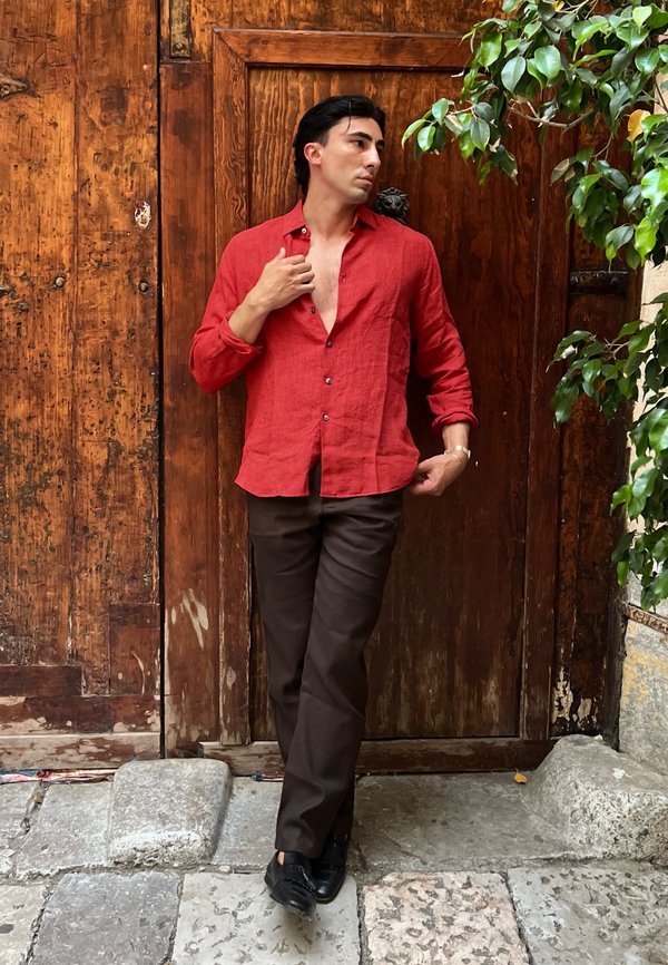 Man in rood overhemd en donkere broek staat ontspannen tegen een houten deur met een groene bladplant aan de rechterkant.
