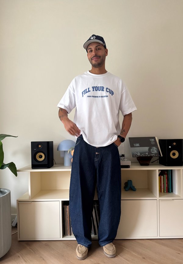 Egy férfi fehér "FILL YOUR CUP" feliratú pólóban, bő sötét farmerben, bézs cipőben és fekete sapkában, aki egy polc mellett áll, amin hangszórók és könyvek vannak.