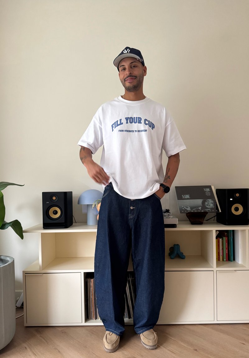 Un homme portant un t-shirt blanc avec l'inscription "REMPLIS TA TASSE", un jean ample foncé, des chaussures beiges et une casquette noire, debout près d'une étagère avec des enceintes et des livres.