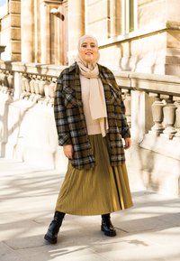 Casaco oversized xadrez em tons escuros, suéter bege, saia plissada verde-oliva e botas pretas com atacadores. Tecidos suaves e texturizados com um look em camadas.