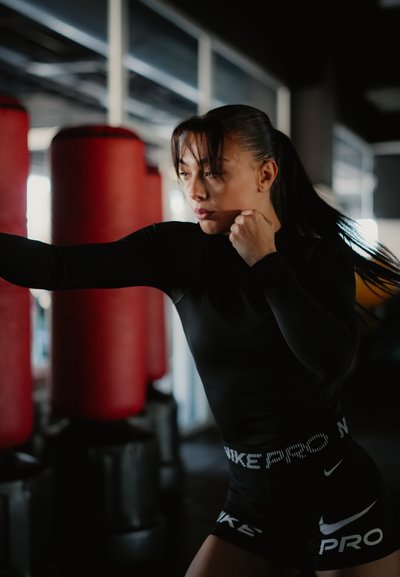Mujer vestida con ropa deportiva negra practicando boxeo con los puños levantados cerca de sacos de boxeo rojos en un gimnasio con poca iluminación.