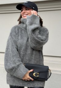 A woman smiling in a black cap and an oversized grey fuzzy sweater holds a black leather clutch with a gold clasp, standing against a beige wall.
