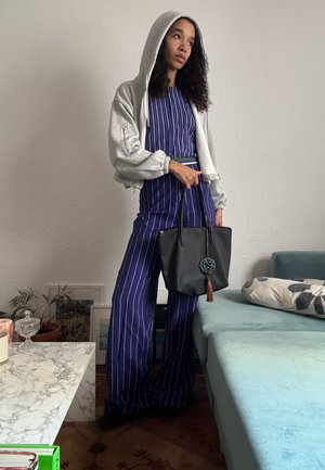 Mujer con pantalones anchos de rayas azules y blusa, con chaqueta con capucha gris claro, sosteniendo una bolsa tote negra, de pie junto a un sofá turquesa.