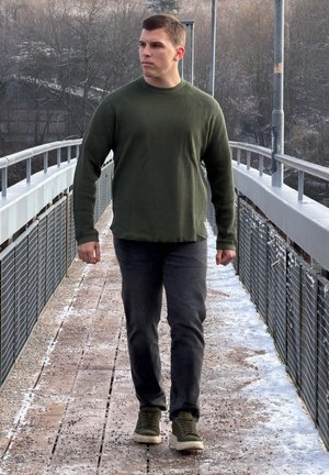 Jersey verde con ribetes, vaqueros gris oscuro y zapatillas oliva. De pie sobre una pasarela de madera, con fondo de árboles y una barandilla.