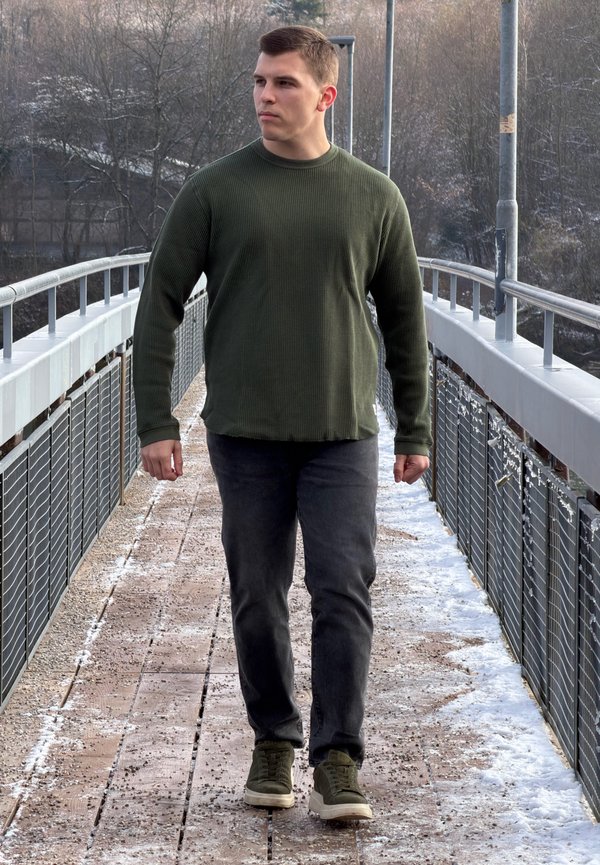Grüner Rippstrickpullover, dunkelgraue Jeans und olivgrüne Sneaker. Steht auf einem Holzsteg, mit Bäumen und einem Geländer im Hintergrund.
