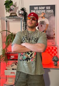 Jeune homme portant une casquette rouge, des lunettes de soleil et un t-shirt graphique, debout les bras croisés dans une pièce avec des plantes, des boules disco, des baskets et une affiche motivante.