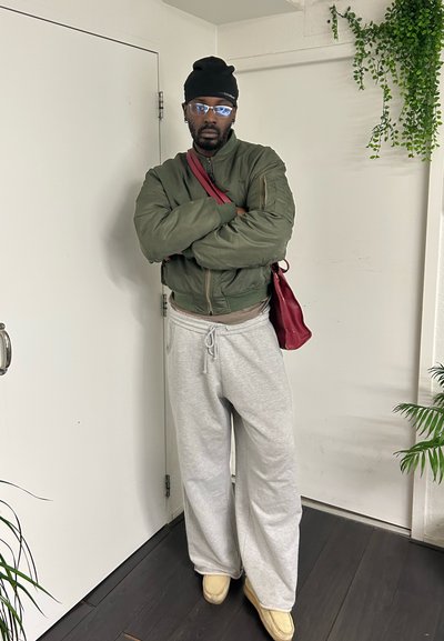 Hombre de pie en el interior con los brazos cruzados, vestido con una chaqueta bomber verde, pantalones deportivos grises, gorro negro, gafas y llevando un bolso rojo de hombro.