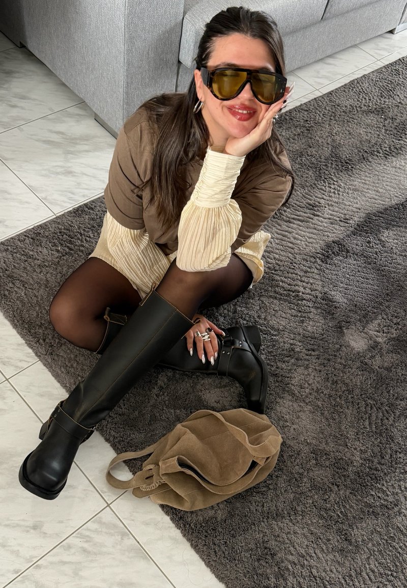 Ženska z dolgimi temnimi lasmi, ki nosi večje sončna očala, rjavo majico, kremne plisirane rokave, črne nogavice in škornje do kolen, sedi na sivi preprogi.